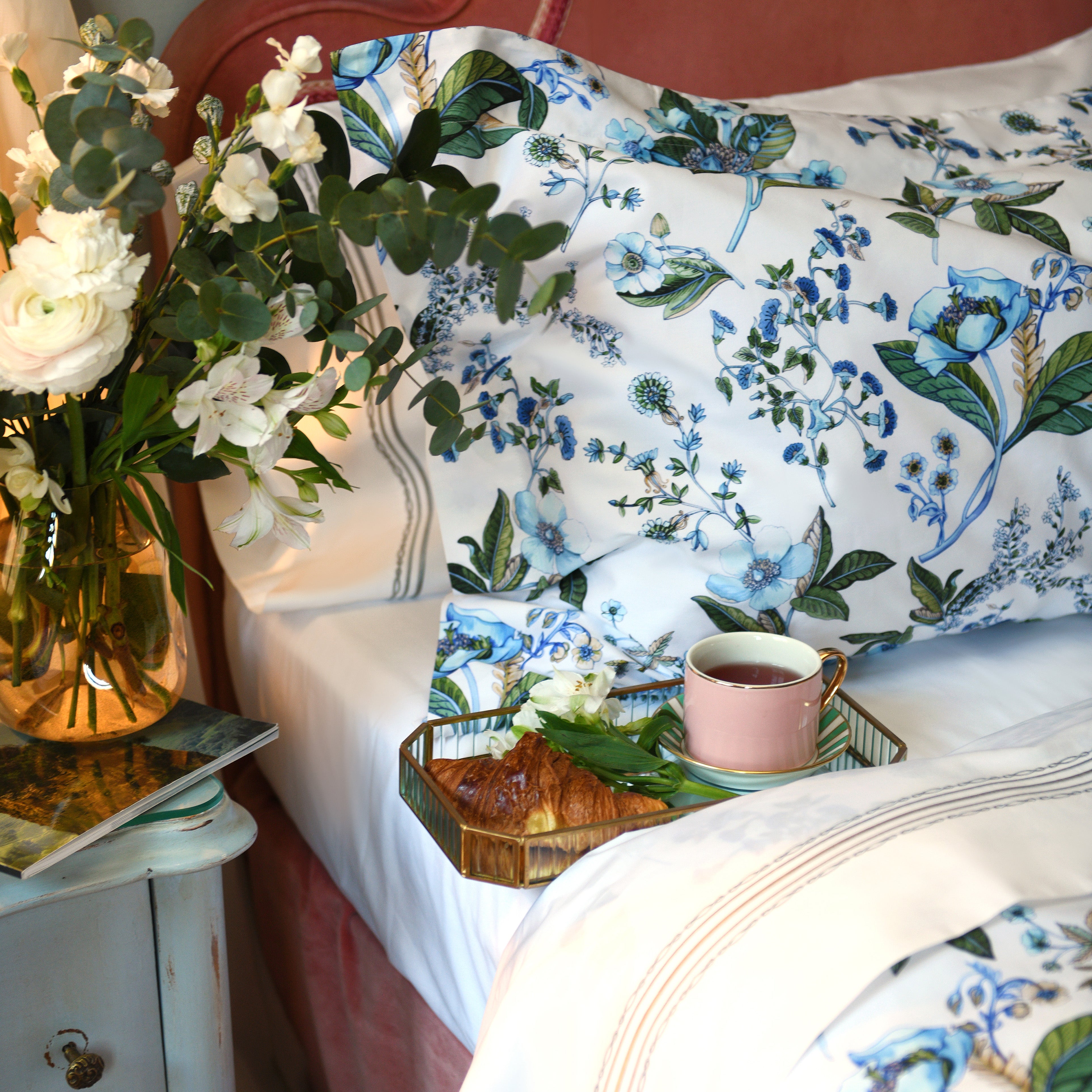 Close-up of pillowcases with Fantasia floral print, croissant and tea on bed with white cotton sheet