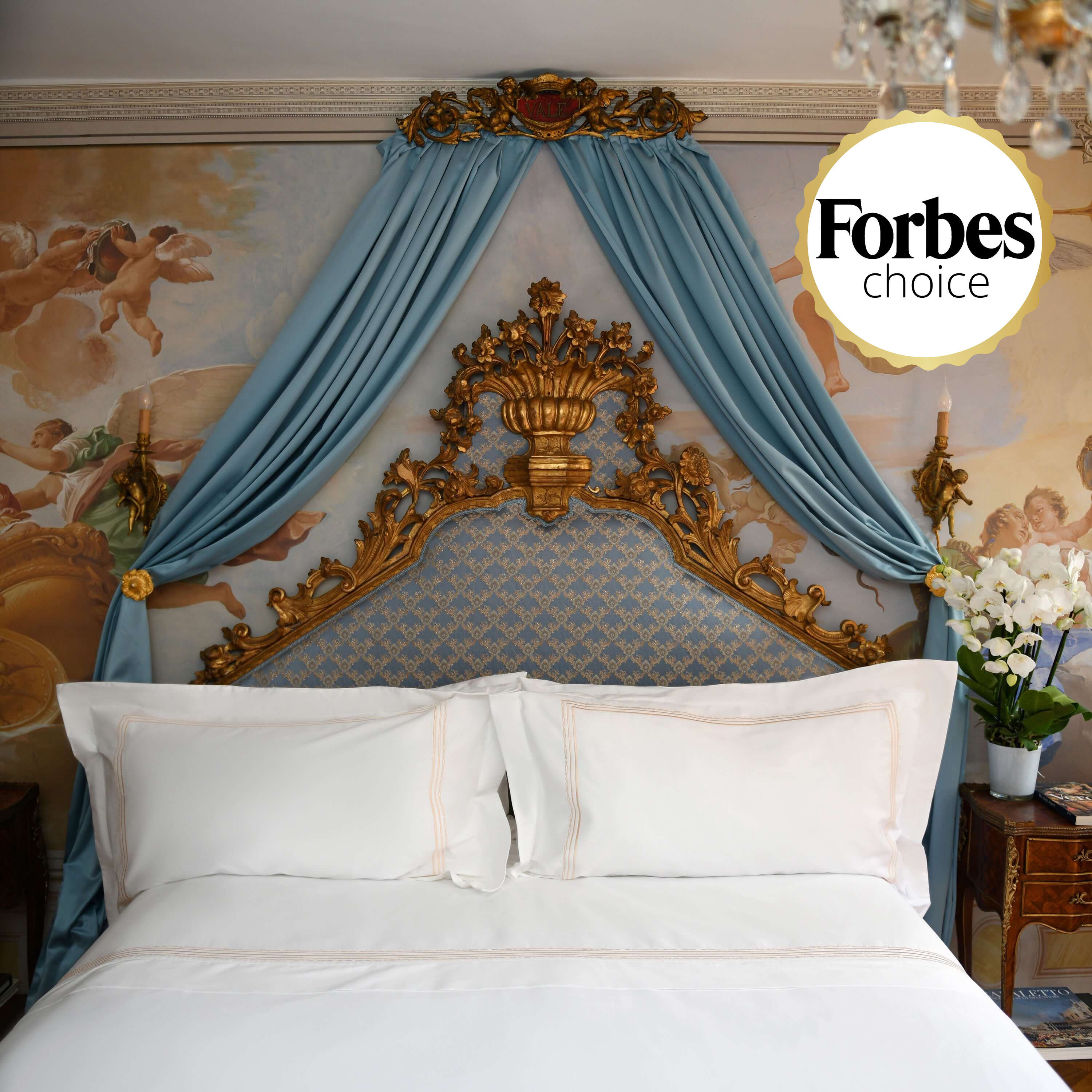 Luxurious white duvet cover set with golden embroidery on regal bed with blue drapery, featured in Forbes