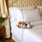 Close-up of Oxford shams with silver embroidery on white cotton bed, with coffee tray and croissants