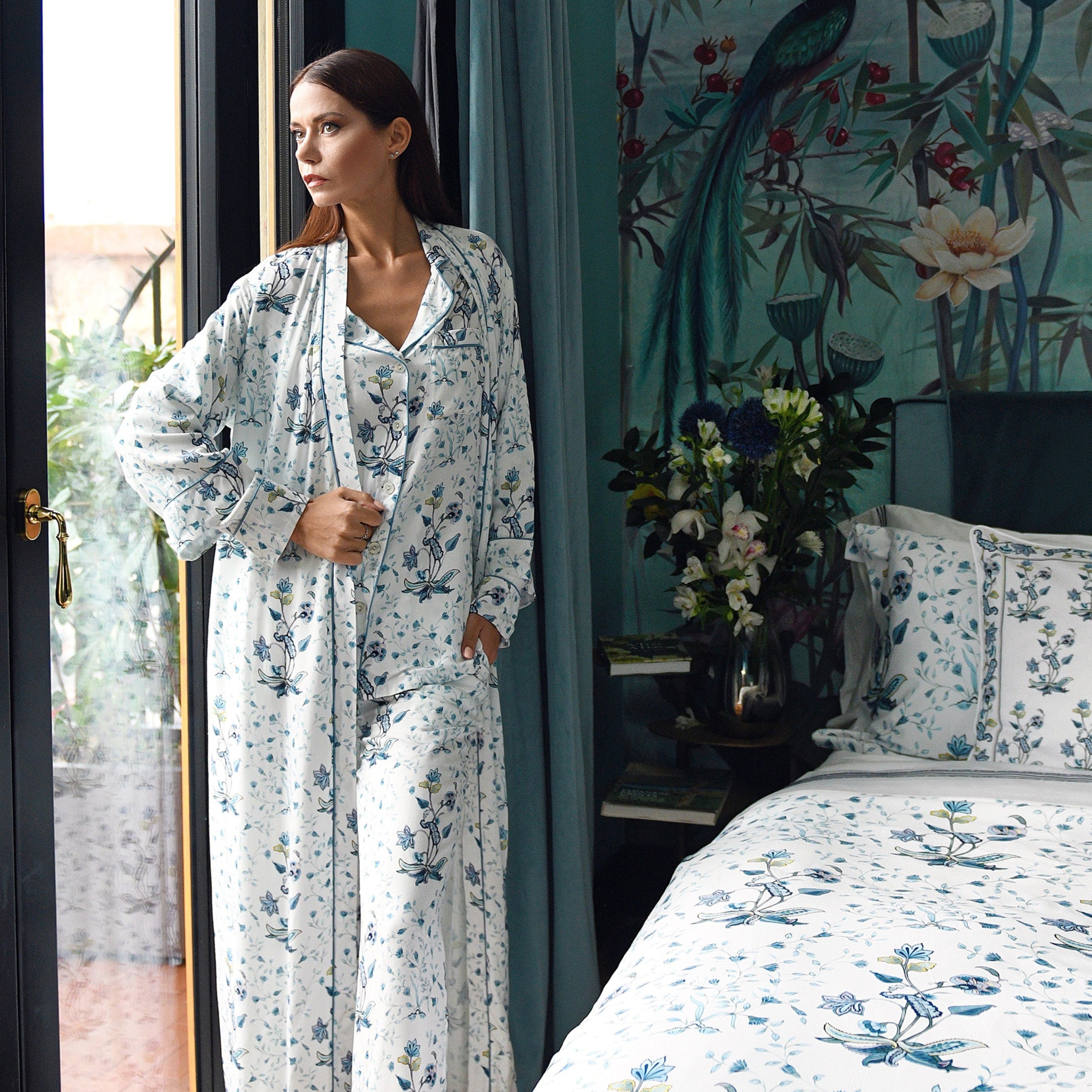 Model in long floral robe standing by window next to bed with printed Fiori cotton bedding