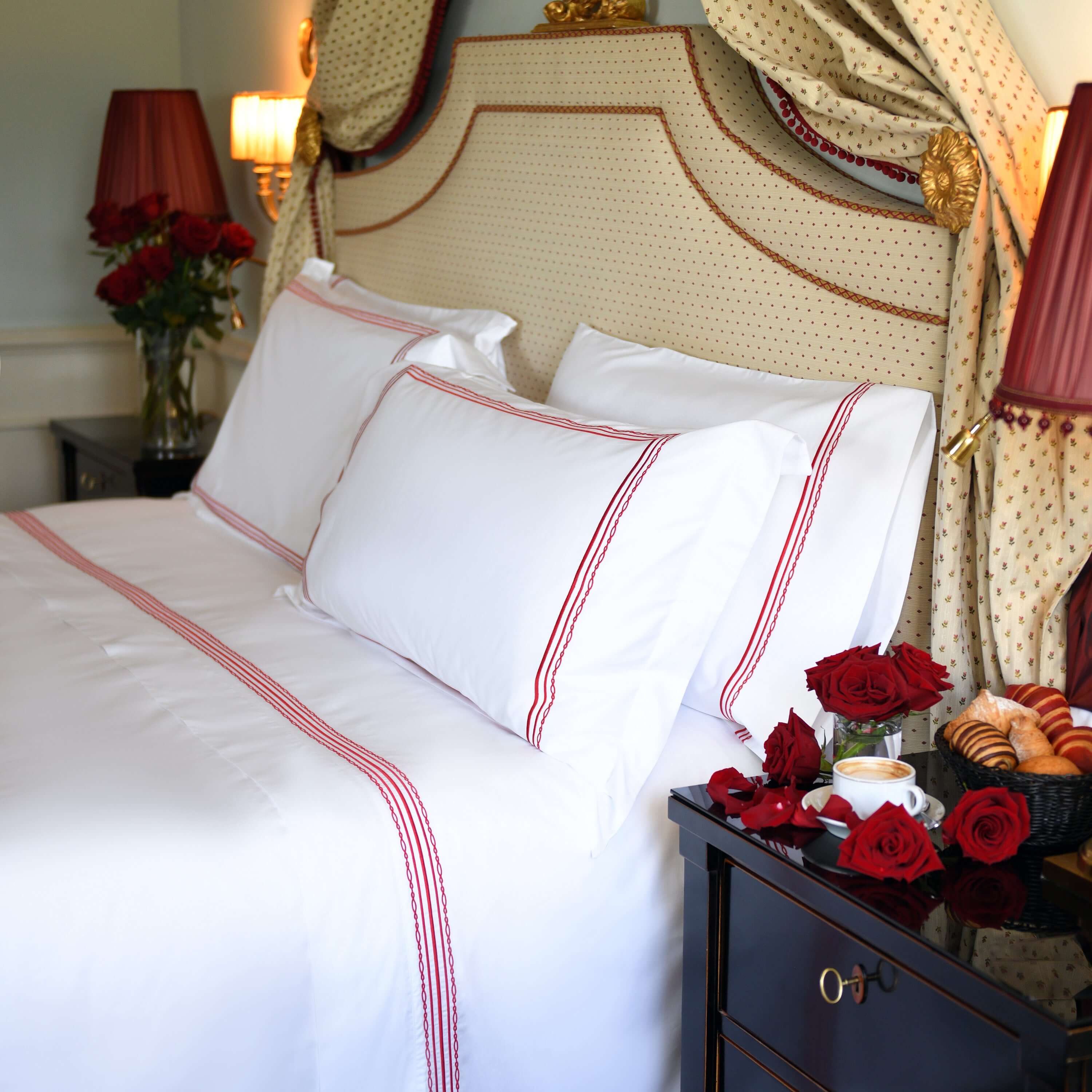 Side view of white cotton duvet with ruby red embroidery, croissants, coffee and red roses on nightstand