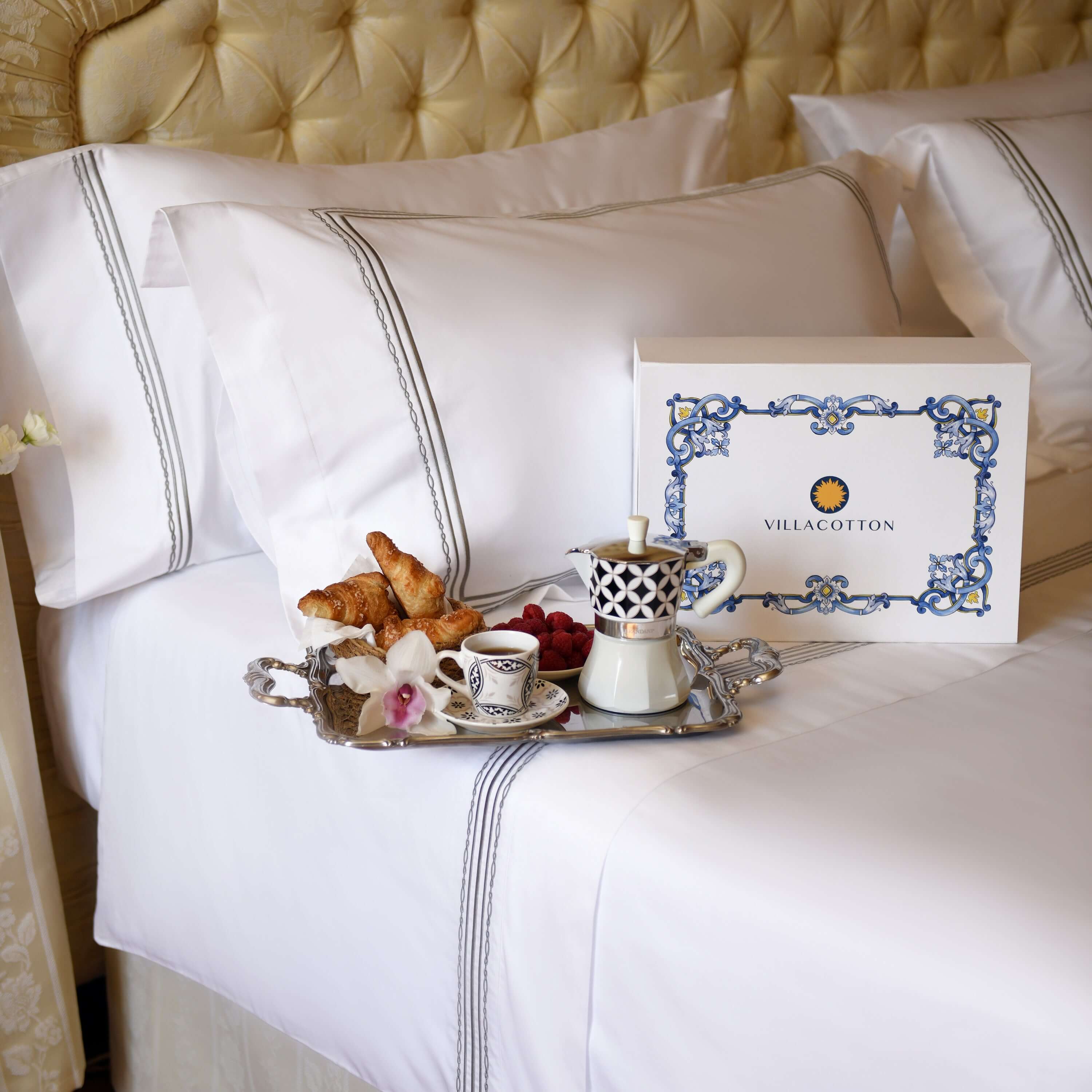 Villa Cotton branded box on white bed with silver embroidered duvet and breakfast tray